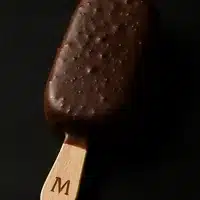 Plaisir glacé : bâtonnet Magnum enrobé de chocolat craquant, tenu par un manche en bois marqué d'un 'M', sur fond sombre.