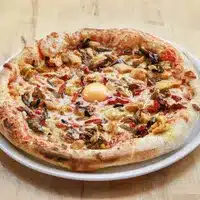 Pizza appétissante à croûte épaisse, garnie de champignons, de morceaux de viande et d'un œuf au centre, servie sur une assiette blanche.