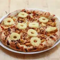 Pizza hawaïenne cuite au four sur plat blanc, garnie d'ananas en rondelles, poulet et fromage fondu, croûte dorée et croustillante.