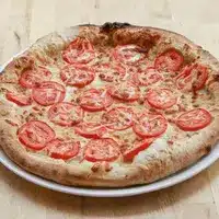 Pizza artisanale cuite au four avec une croûte épaisse et dorée, généreusement garnie de tranches de tomate fraîche et de fromage fondu, présentée sur une assiette blanche.