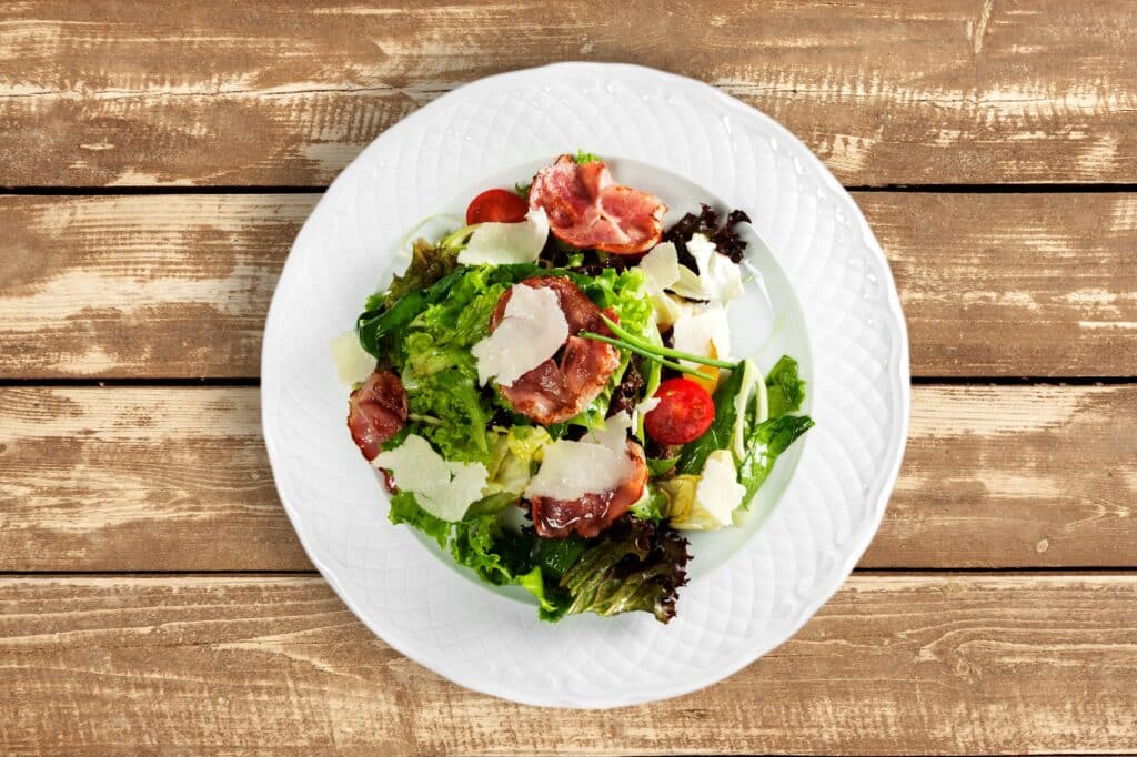 Salade fraîche avec lardons croustillants, copeaux de parmesan et tomates cerises, servie sur une assiette blanche sur fond de bois rustique.