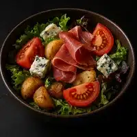 Salade gourmande avec pommes de terre rôties, tranches de tomates, dés de fromage bleu persillé, et fines tranches de jambon cru sur un lit de laitue verte.