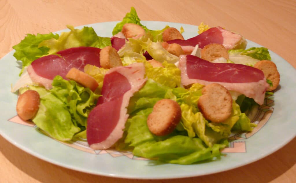 Salade composée de laitue verte et jaune, garnie de fines tranches de magret de canard séché et de croûtons ronds, servie sur une assiette claire.