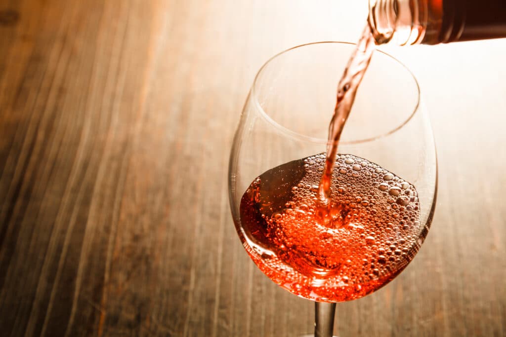 Verseur de vin rosé pétillant dans un verre, créant une mousse de bulles sur fond de bois sombre.